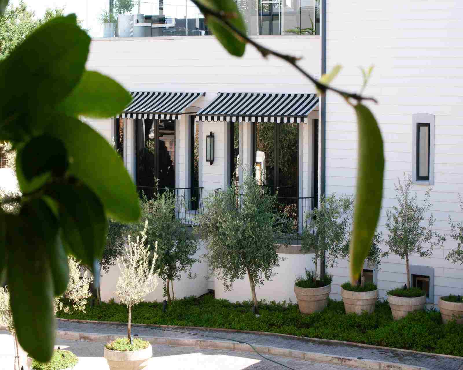 Exterior view of a white, timber hotel building with black-and-white awnings and iron balconies with trees and potted plants outside.