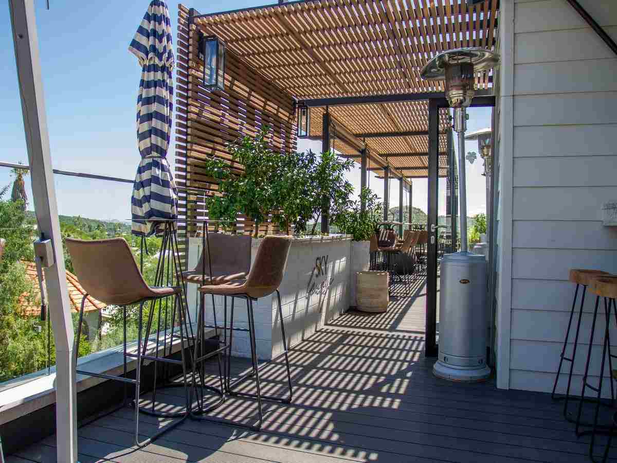 A rooftop terrace with pergola, bar stools, patio heaters and striped parasols, with views of greenery visible all around.