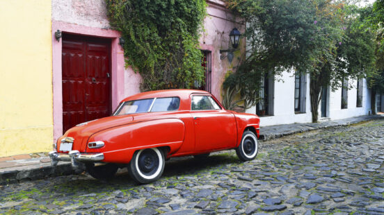 Vintage red car parked on a cobblestone road beside a pink building and green climbing vines.