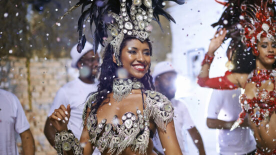 A woman in a sparkling carnival costume and feathered headpiece smiles as confetti falls around her.