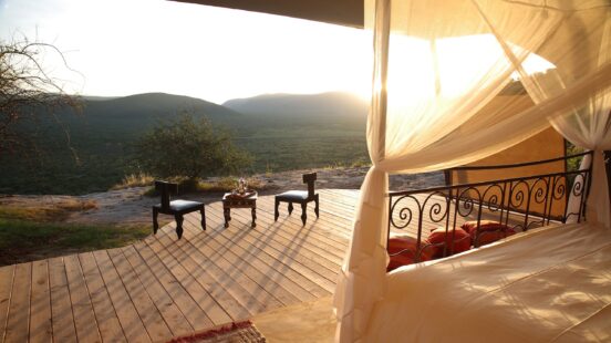 View from inside a luxury tent with a canopy bed looking out to a wooden deck with two chairs overlooking a green valley.
