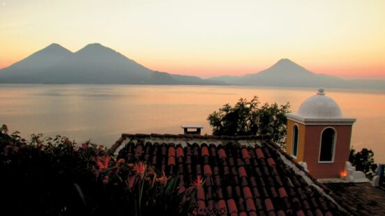 View from a tiled roof toward a calm lake and two large volcanoes under a soft orange sunset sky.