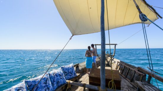 A man and woman stand on the deck of a wooden dhow sailboat, looking at the horizon on a sunny day.