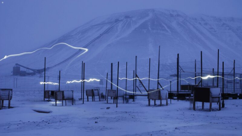 A snowy field with wooden huts and white light streaks in front of a massive, dim mountain during blue hour.