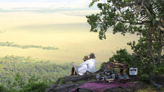 Two people sitting on a cliff overlooking a vast savanna, with a picnic basket and blanket nearby.