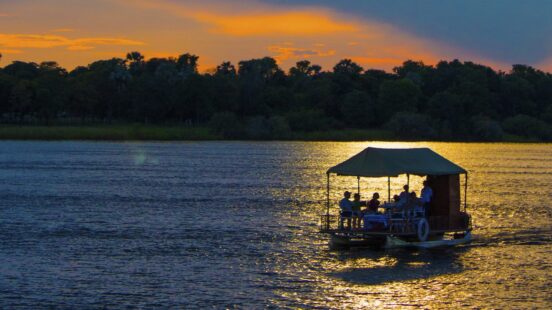 Silhouetted passengers on a covered boat cruising along a river at sunset with golden light reflecting on the water.