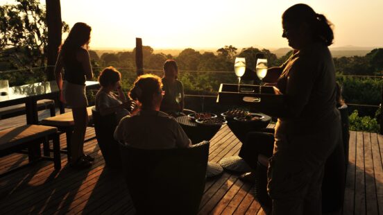 Silhouette of a server holding a tray of glasses for guests on a deck at sunset.