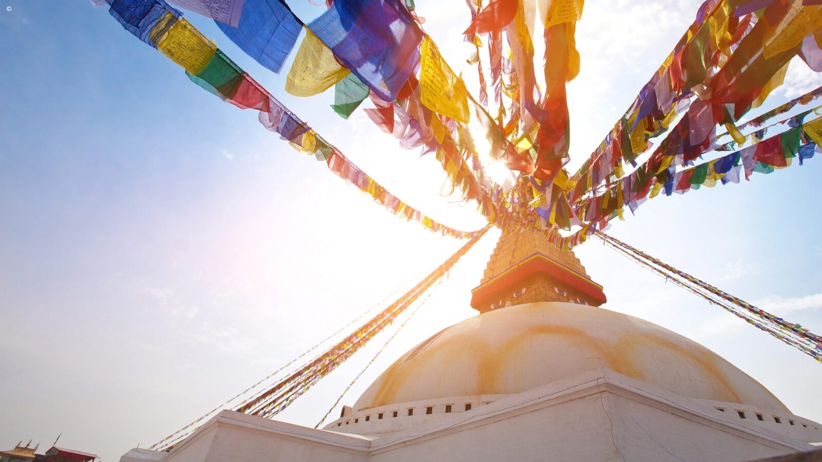 Sunlight shines through rows of colorful Buddhist prayer flags connected to the golden spire of a white domed stupa.