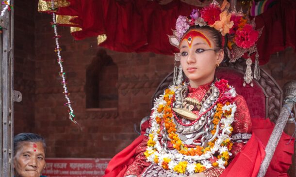 A young girl dressed as the Kumari in ornate traditional red clothing and heavy silver jewelry sits on a decorative throne.