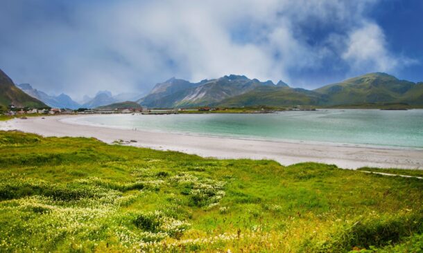 A sweeping view of a sandy beach, green meadows, and jagged mountains under a blue and cloudy sky.