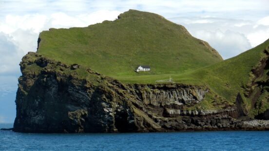 A small white house standing alone on a bright green grassy cliffside of a mountainous island.
