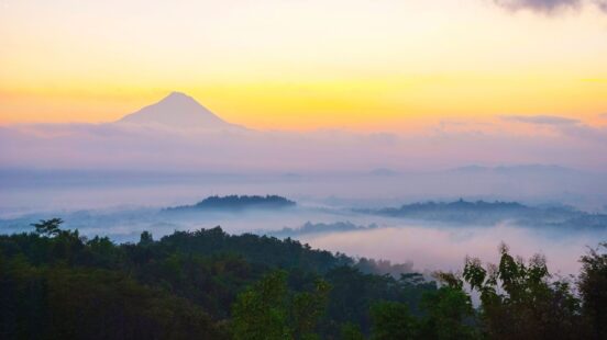 Misty mountain peak rising above a layer of clouds and forest during a colorful sunrise with orange and blue hues.