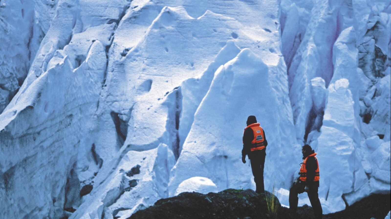 A low-angle view of two individuals in dark winter clothing and bright orange life vests standing before a towering, craggy white and blue ice glacier.