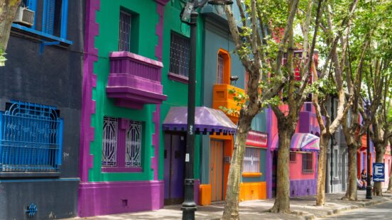 A row of colorful houses, painted in bright purple, green, orange, and blue, line a sunlit street.