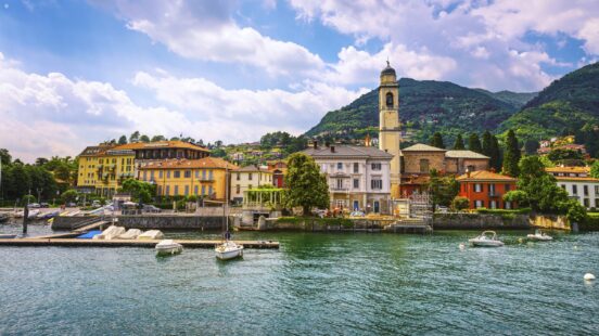Waterfront view of an Italian village with a prominent stone bell tower against a mountain backdrop.