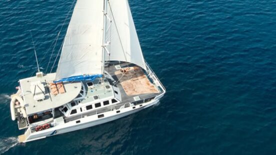 Aerial view of a white sailboat cruising on clear blue ocean water with a large white sail.