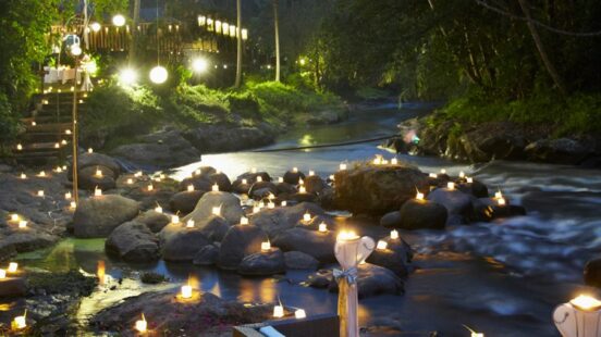 Many glowing candles placed on rocks in a river at night, creating a romantic atmosphere in a lush forest.