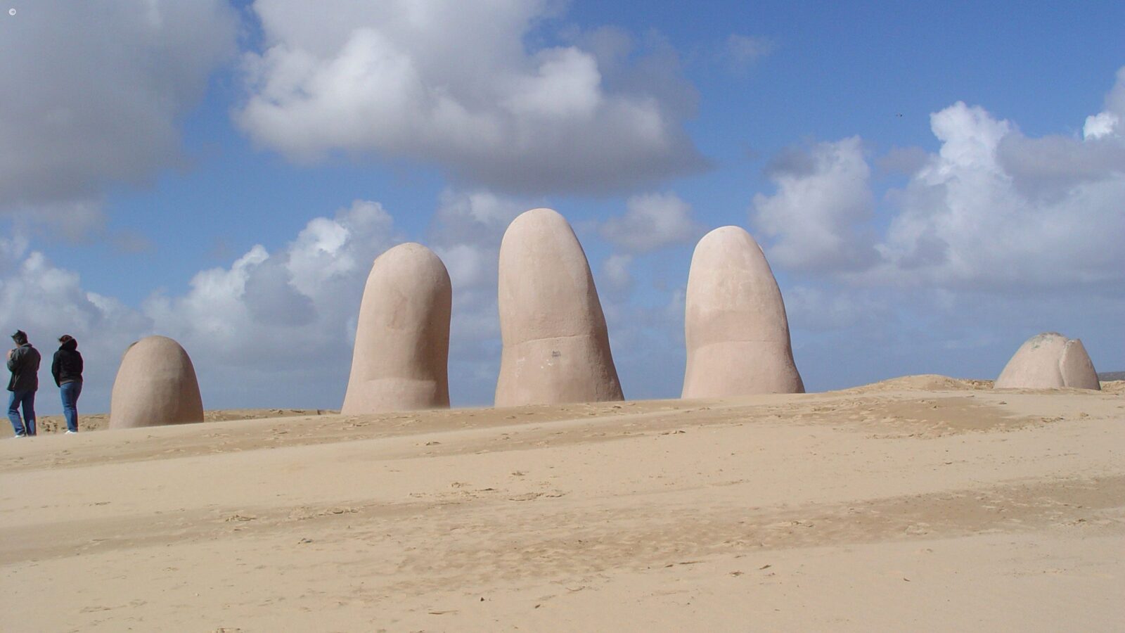 Large stone sculpture of a hand rising from the sand dunes with people walking nearby under a blue sky.