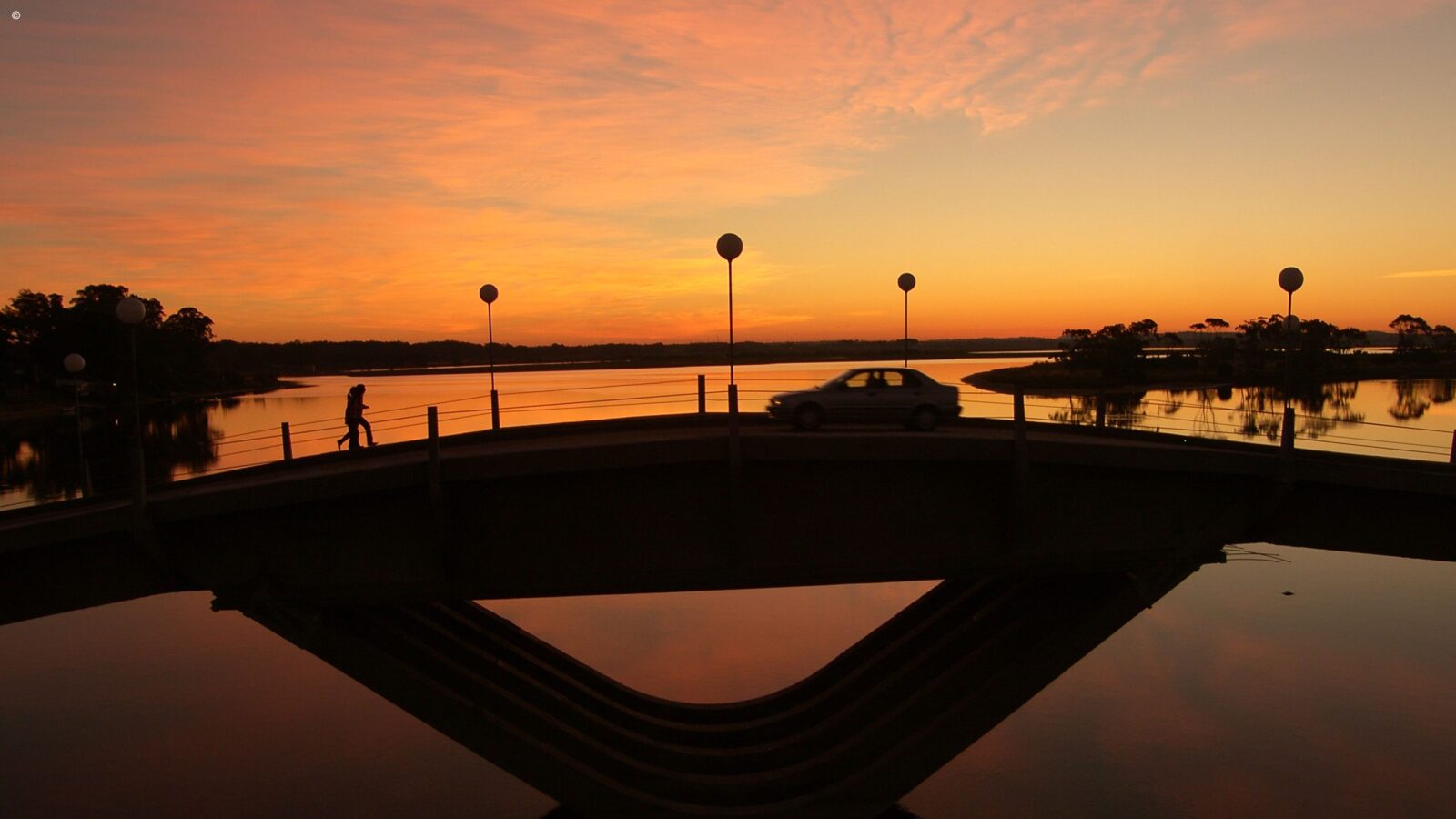 Silhouette of a wavy bridge over water at sunset with a car and a person walking across.