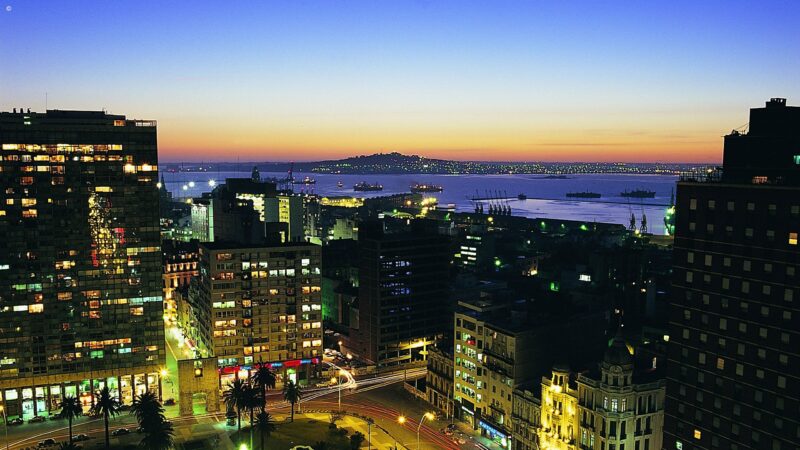 Nighttime aerial view of an illuminated city skyline and a harbor with ships under a dark sunset sky.