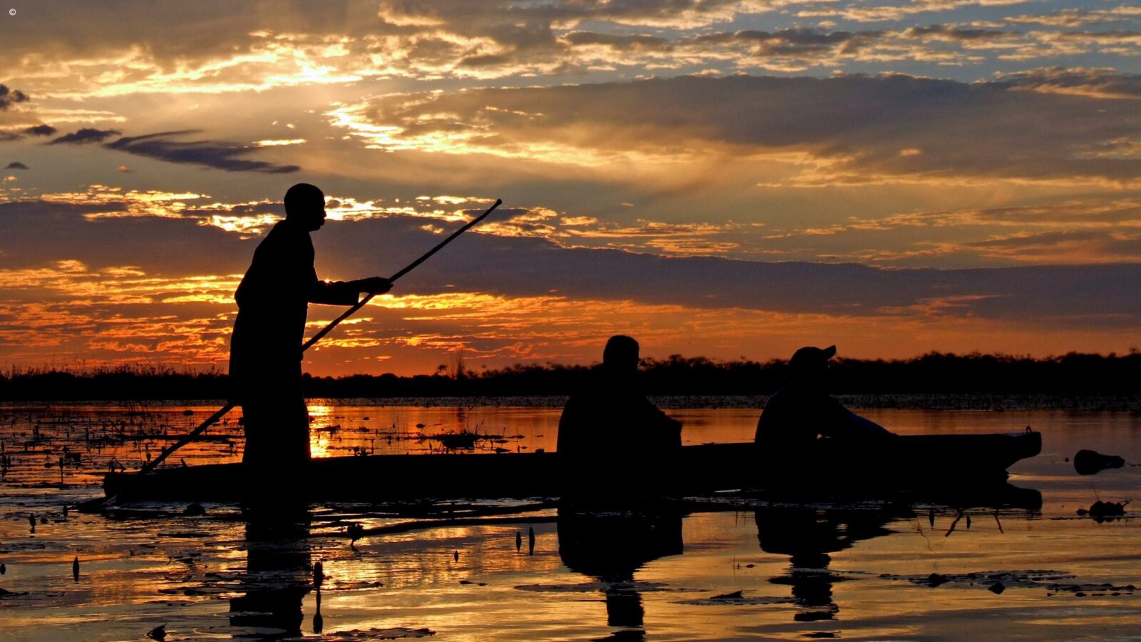 Silhouetted people in a boat at sunset on luxury Okavango Delta Tours.
