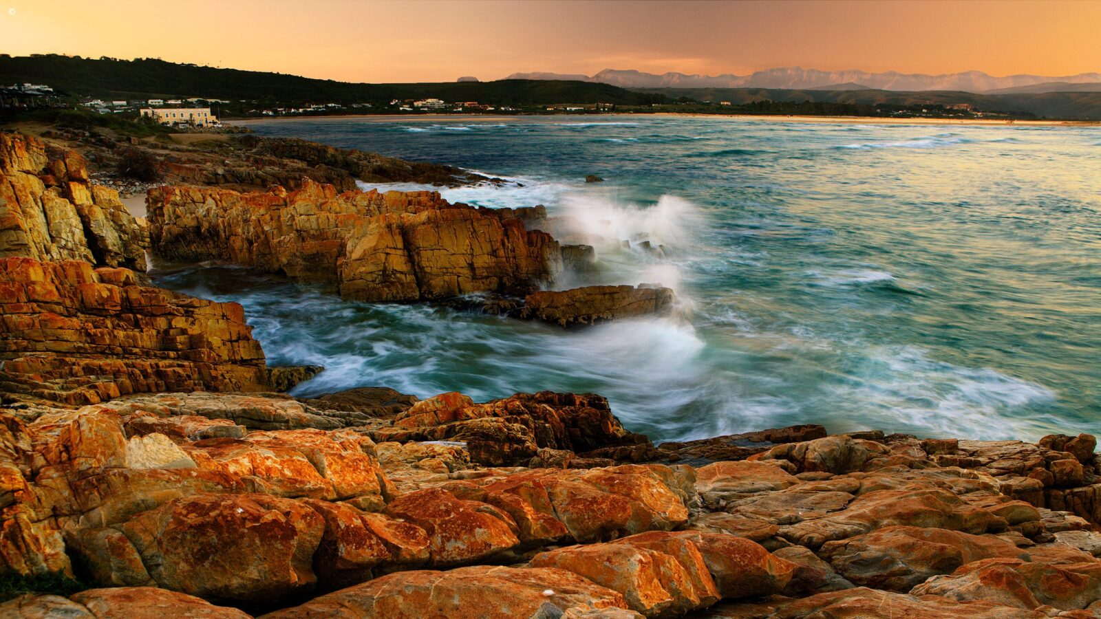 Waves crashing against orange rocks during a luxury garden route tour at sunset with mountains in the background.