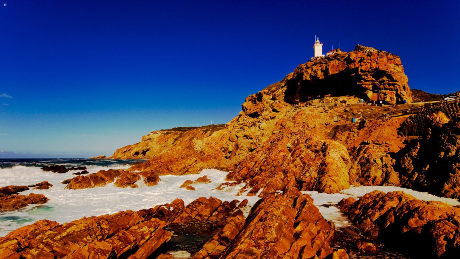 A white lighthouse on a golden rocky cliff above crashing waves, perfect for luxury garden route trips.