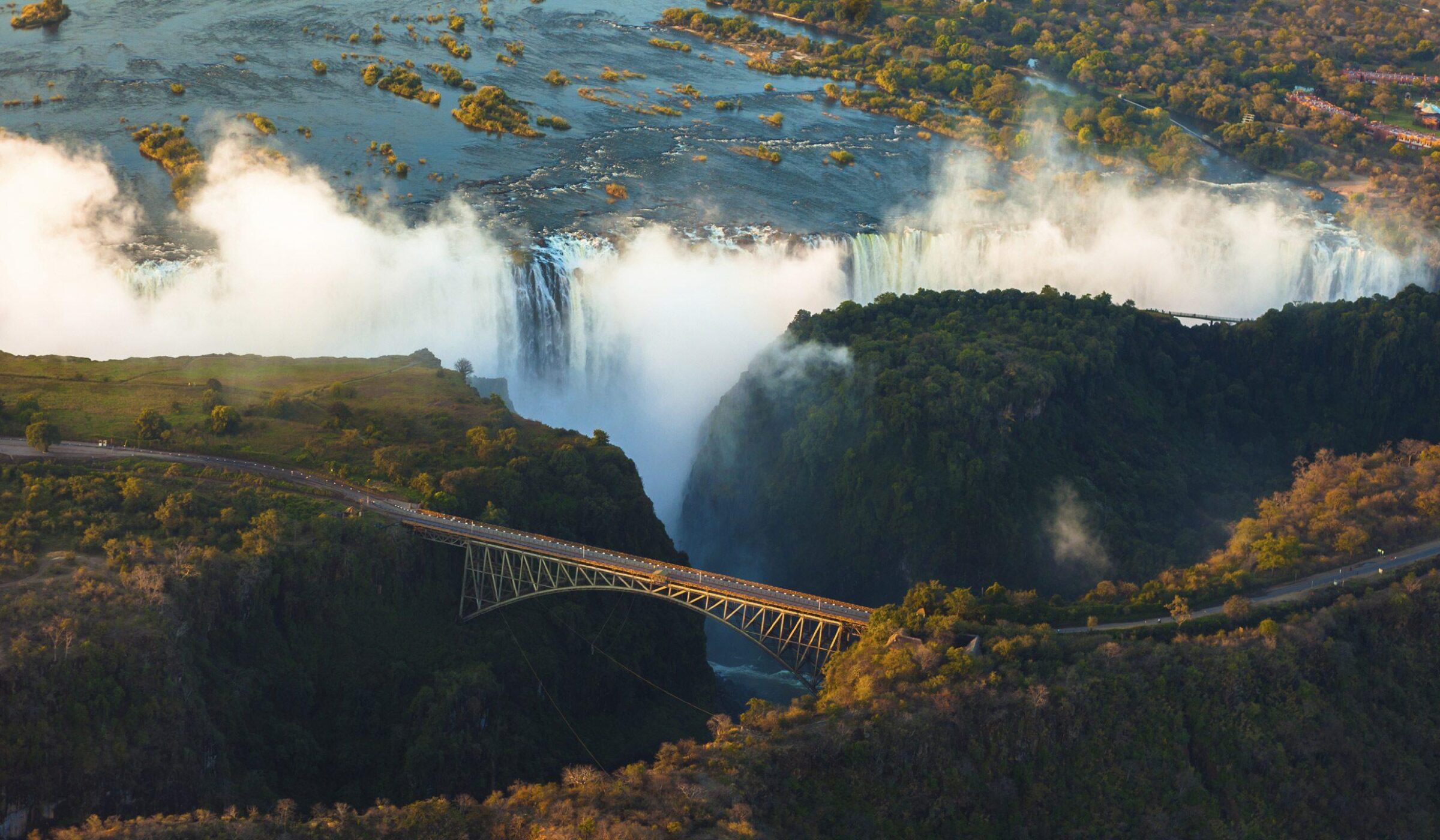 Aerial view of a stunning waterfall surrounded by lush greenery, with a bridge crossing over a gorge below the misty falls.
