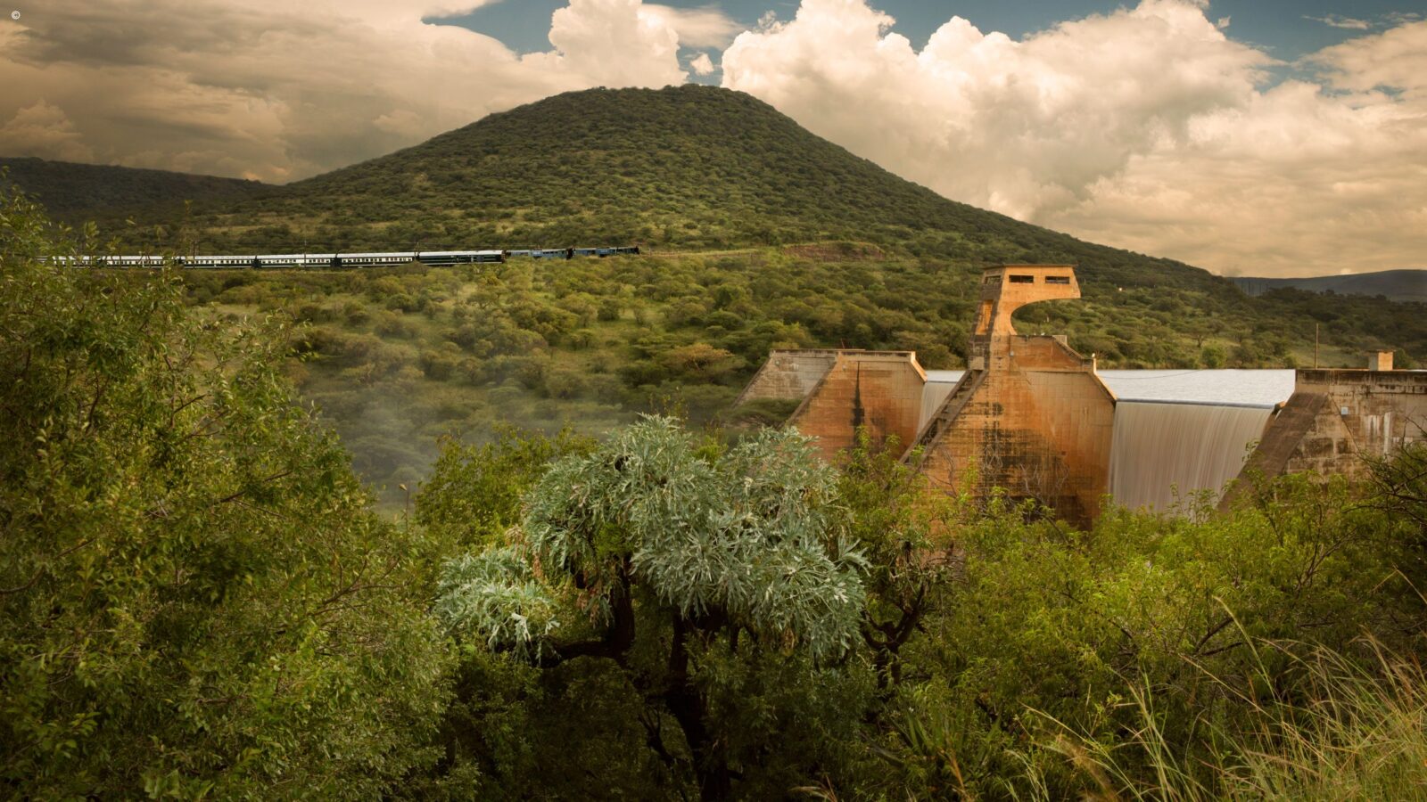 A long train for luxury South Africa rail tours travels through a green valley past a large dam.