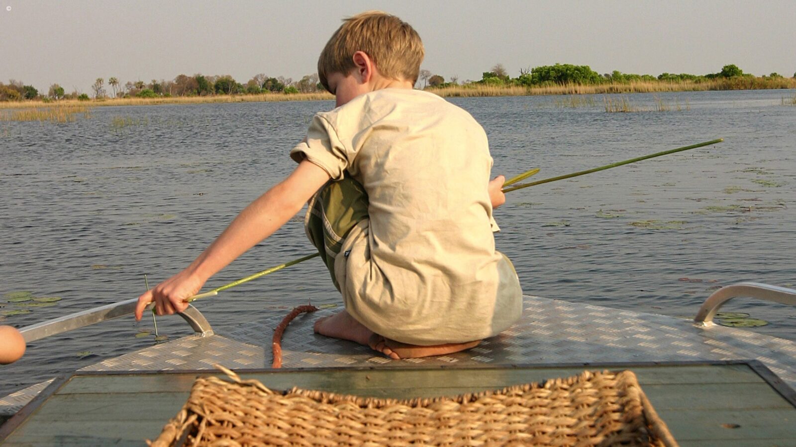 A young boy with blonde hair crouching on the edge of a boat with his back to the camera, holding a long piece of grass