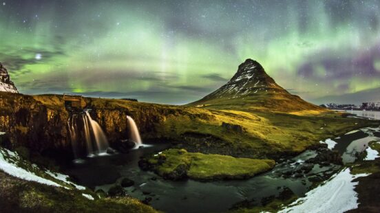 Kirkjufell mountain and Kirkjufellsfoss waterfall are visible at night under the green and purple Northern Lights, inspiring luxury Iceland vacations.