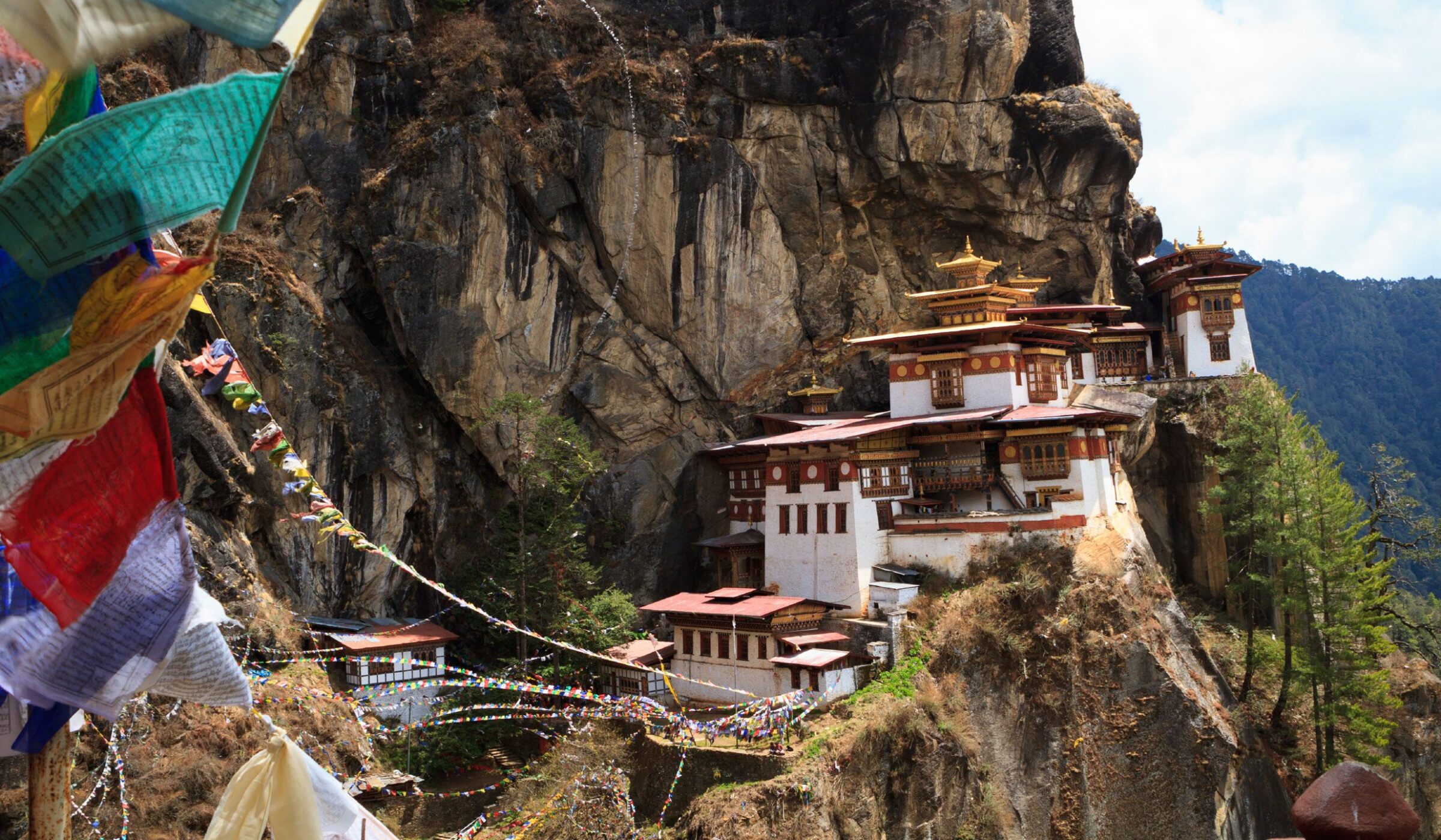 A white monastery with golden roofs built into a sheer rock cliff, viewed through hanging prayer flags.