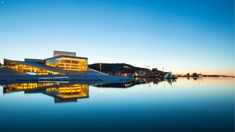 A modern architectural building with glowing interior lights reflecting on still water under a clear dusk sky.