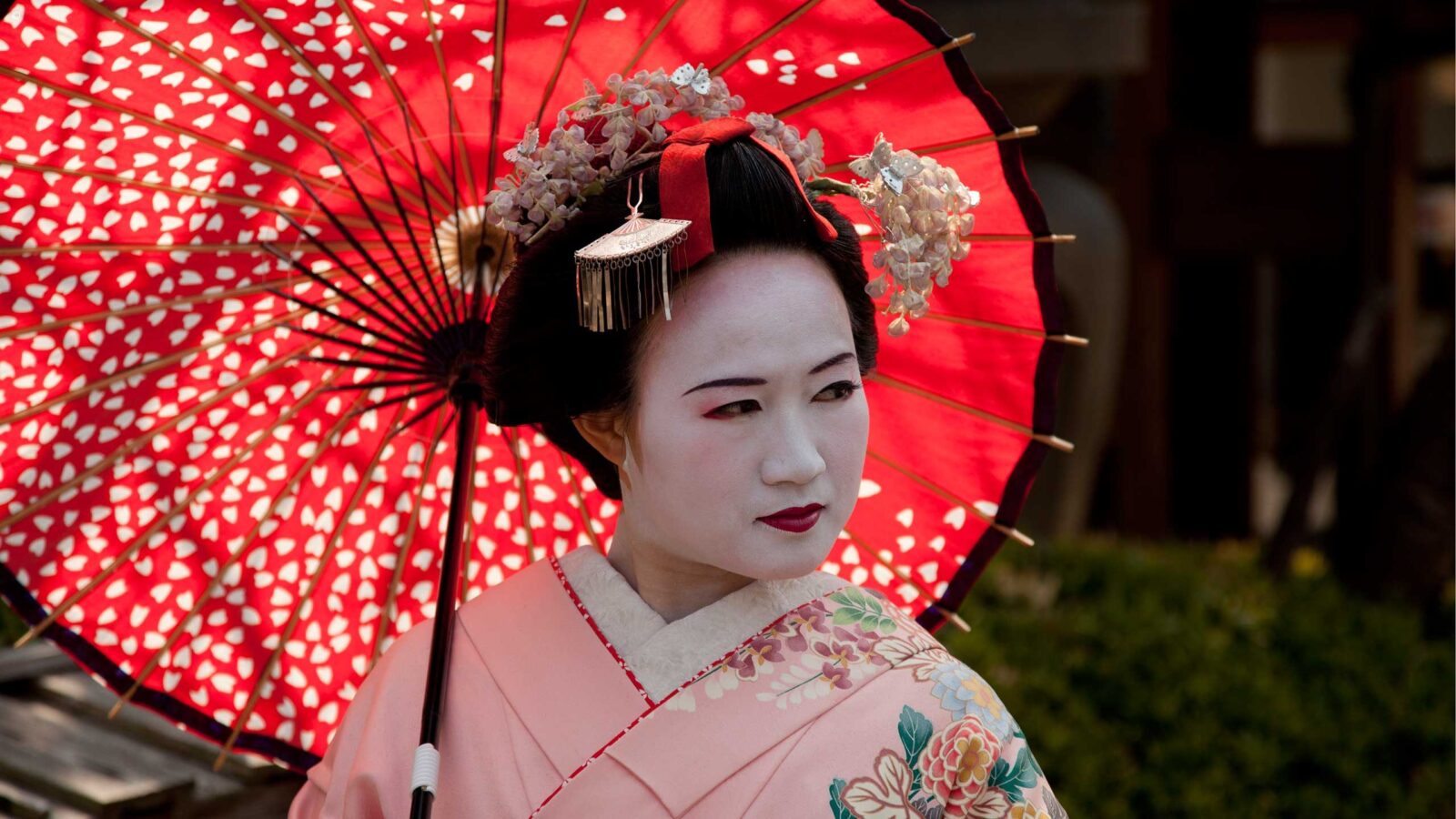 Side view of a woman in a traditional kimono and geisha makeup walking past historic wooden buildings.