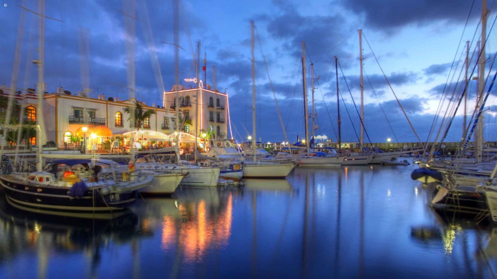 Sailboats docked in a harbor at dusk with lights from a building reflecting on the calm water.