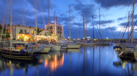 Sailboats docked in a harbor at dusk with lights from a building reflecting on the calm water.