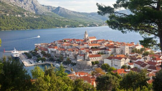 A dense coastal village with red-roofed houses situated on a peninsula against a mountainous backdrop.