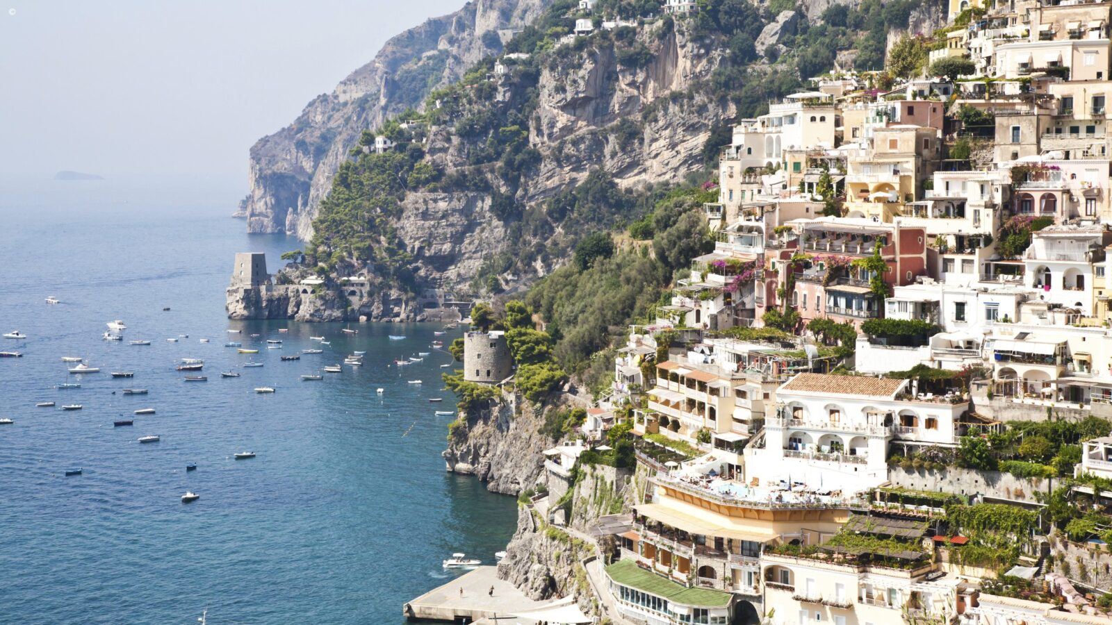 Pastel houses stacked on a steep hillside overlook a sea dotted with white boats on luxury Amalfi Coast trips.