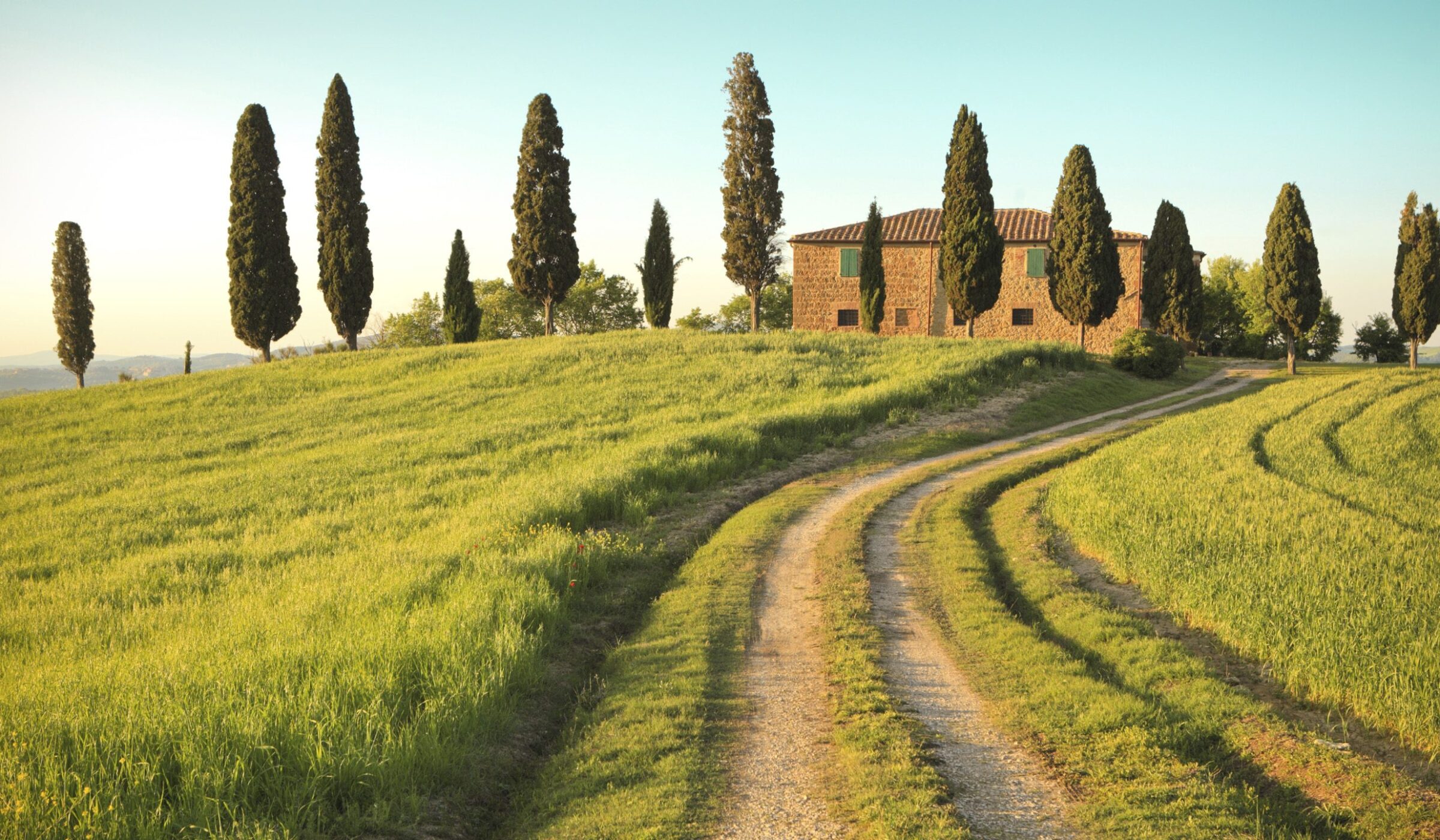 A stone farmhouse on a grassy hill with tall cypress trees and a dirt path, ideal for luxury Tuscany tours.