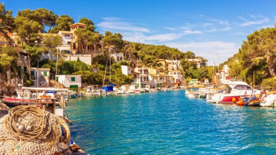 Boats moored in a bright blue harbor surrounded by sunlit buildings and green forest.