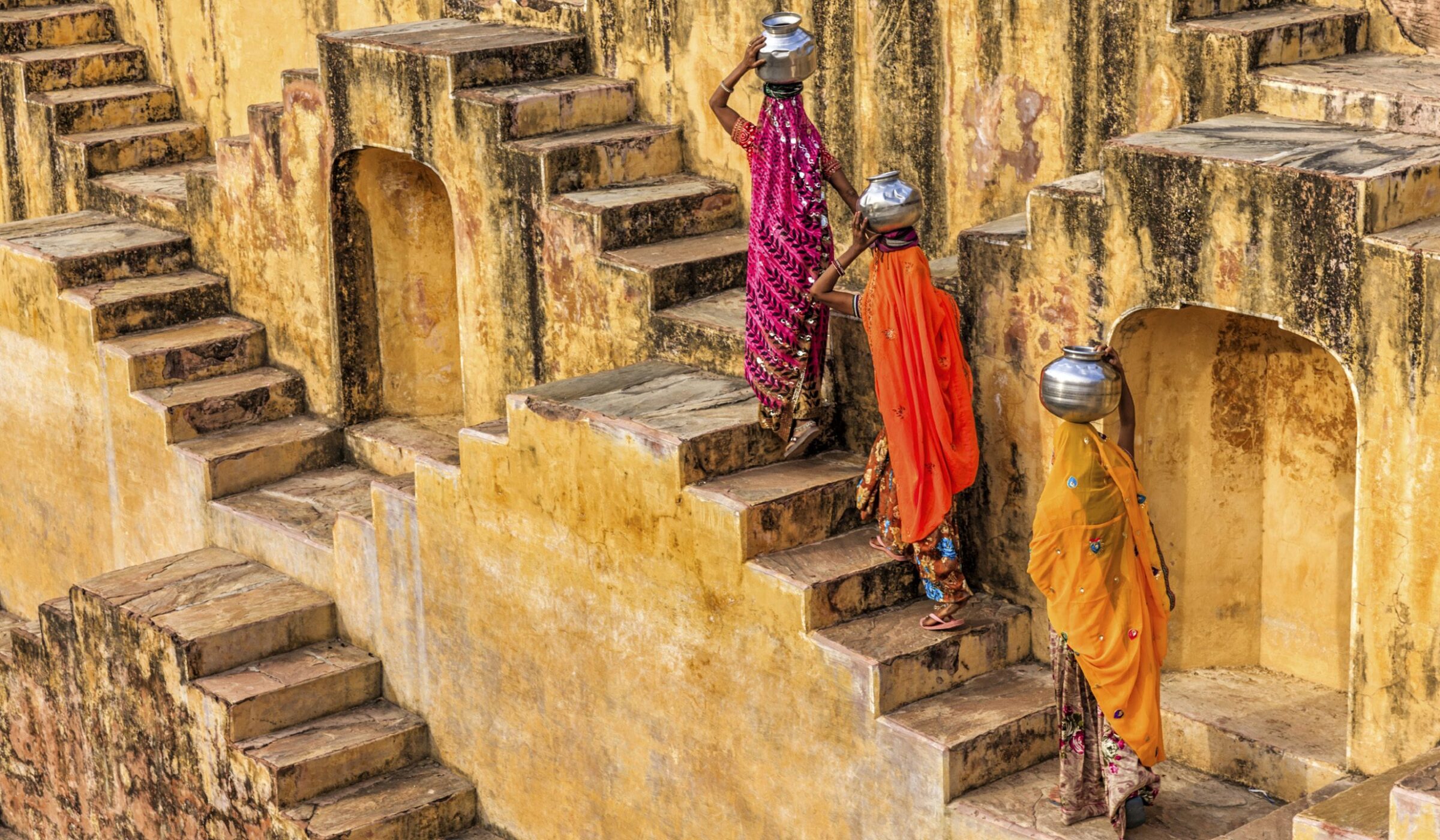 Three women in colorful saris carrying water pots on their heads ascend the geometric steps of a stone stepwell on luxury India trips.