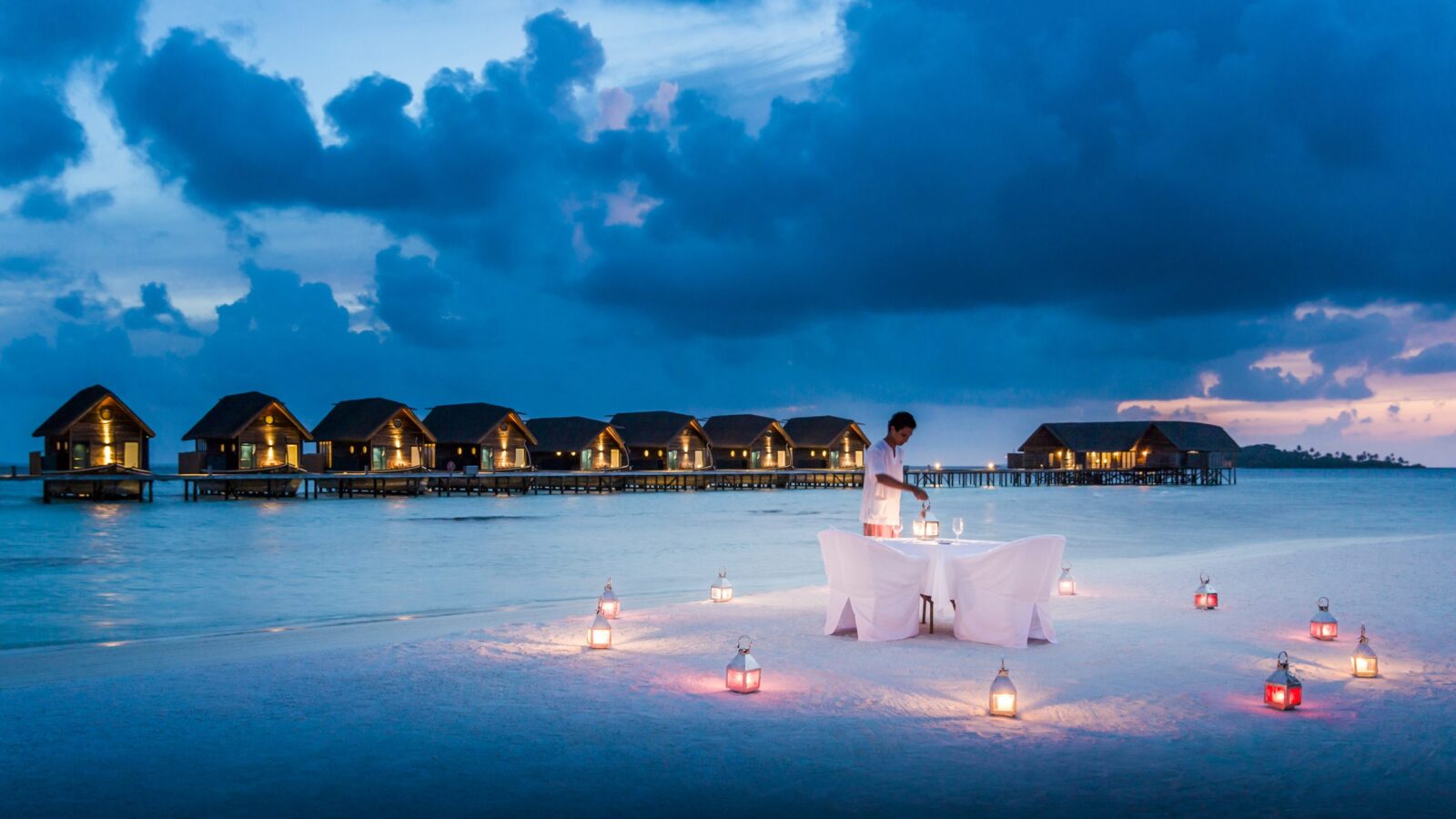 A man setting up a romantic candlelit dinner on a white sandy beach with overwater bungalows in the background