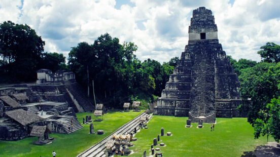 Ancient Mayan stone pyramids and ruins in a large green field surrounded by jungle.
