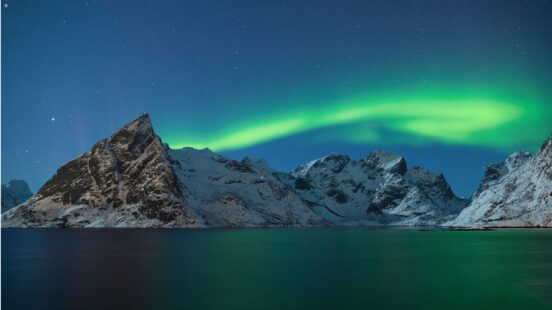 Green northern lights glowing in the night sky above dark water and snowy mountains.