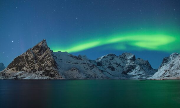 Green northern lights glowing in the night sky above dark water and snowy mountains.
