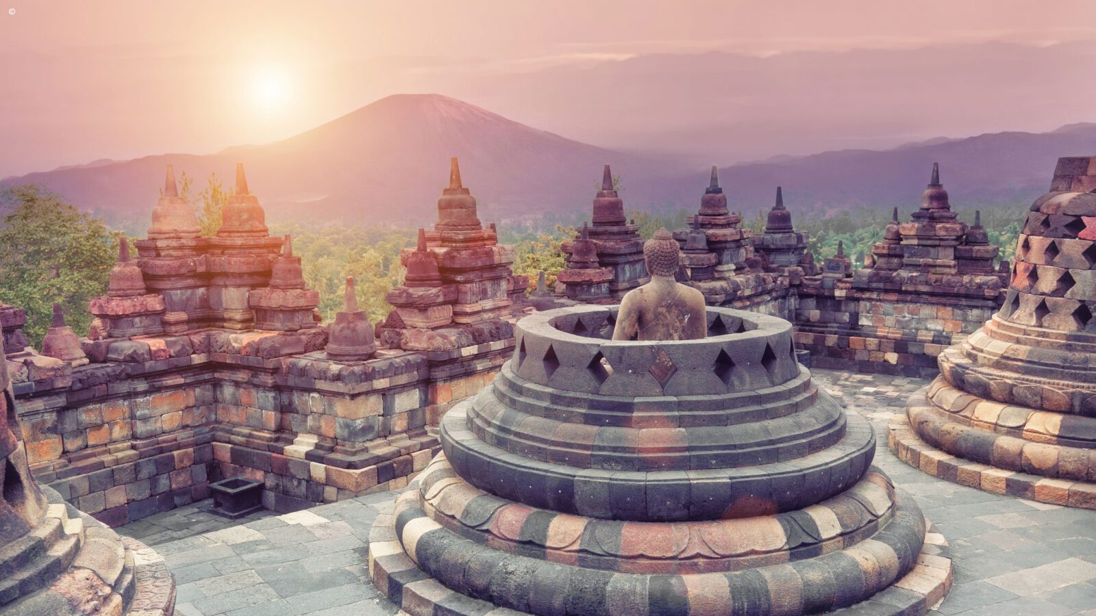 A stone Buddha statue sits among the stupas of Borobudur temple as the sun sets behind mountains for Asia honeymoons.