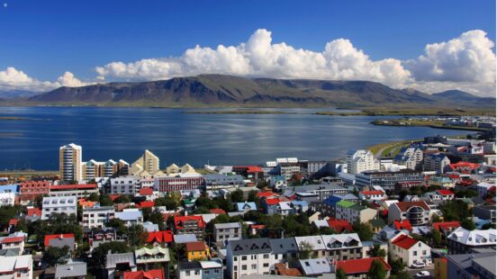 A coastal city with colorful buildings and red roofs set against a large mountain and blue water.