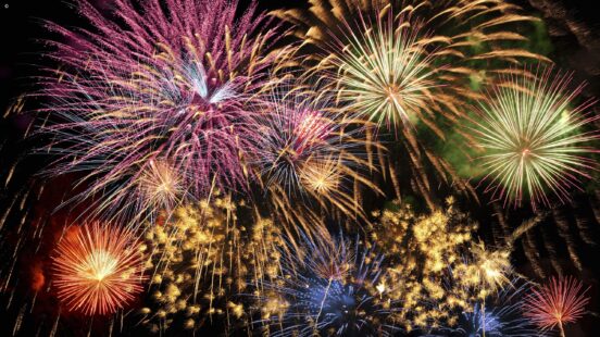 A spectacular display of multiple colorful fireworks exploding in shades of pink, yellow, and blue against a black sky.