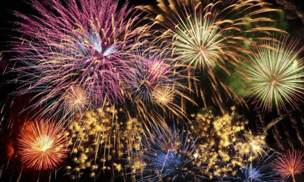 A spectacular display of multiple colorful fireworks exploding in shades of pink, yellow, and blue against a black sky.
