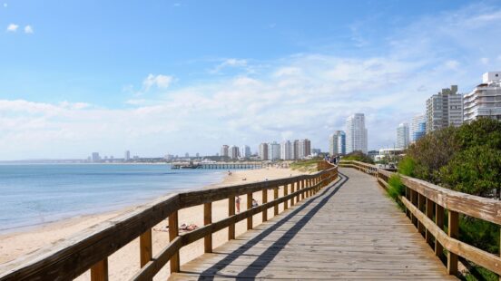 Wooden boardwalk path alongside a sandy beach with high-rise buildings and a pier in the distance.